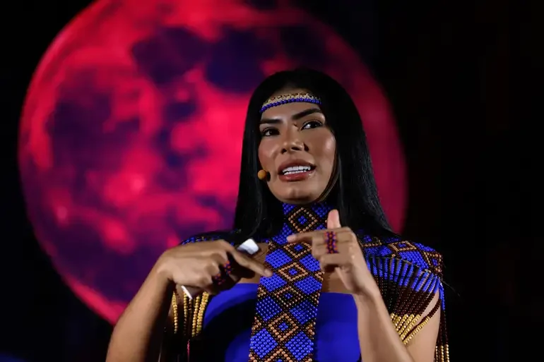 Imagem colorida mostra a cunhã-poranga do Caprichoso Marciele Albuquerque em discusso com roupas e acessórios indígenas azuis e ao fundo uma imagem de um luar avermelhado. Marciele Albuquerque é a dançarina do Caprichoso durante a apresentação da toada Guerreira das Lutas