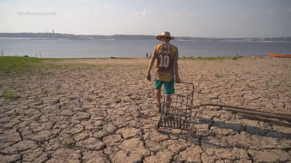 Imagem colorida mostra homem velho de costa arrastando carrinho de compras pequeno com uma mãe e o chão quebrado em virtude de seca de rio, que é visto ao fundo